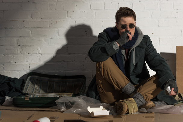 безпритульний чоловік курить сигарету, сидячи на сміттєвому звалищі
 - Фото, зображення