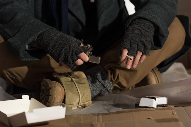 частковий вигляд бездомного чоловіка в рукавичках без пальців, що тримають запальничку та сигарету
 - Фото, зображення