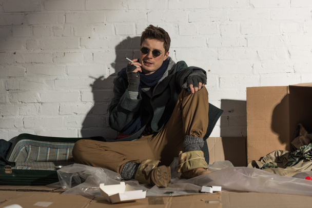 vagabundo sentado en el basurero y fumando cigarrillo
 - Foto, Imagen