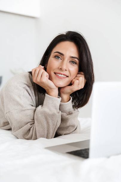 Photo of attractive woman 30s using laptop while lying in bed with white linen in bright room - Photo, Image