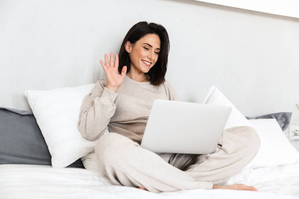 Photo of happy woman 30s using laptop while sitting in bed with white linen in cozy apartment - Fotó, kép