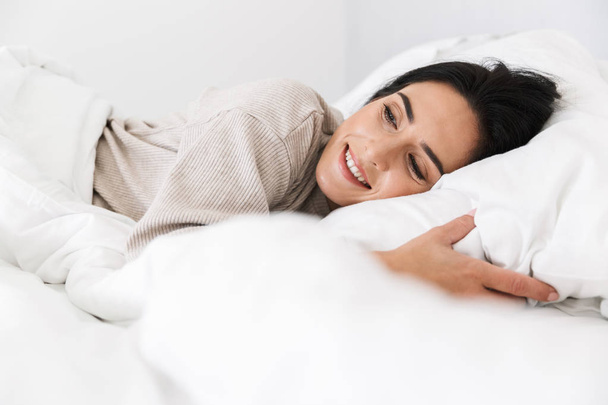 Photo of adorable woman 30s smiling while lying in bed with white linen at home - Foto, afbeelding