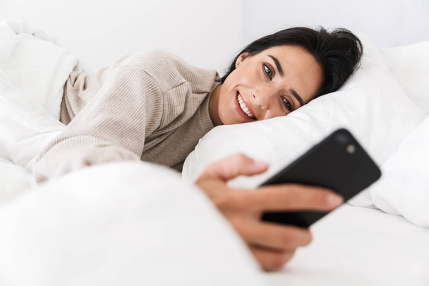 Photo of beautiful woman 30s using smartphone while lying in bed with white linen at home - Photo, image