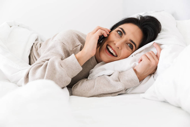 Image of smiling woman 30s using smartphone, while lying in bed with white linen at home - Foto, afbeelding