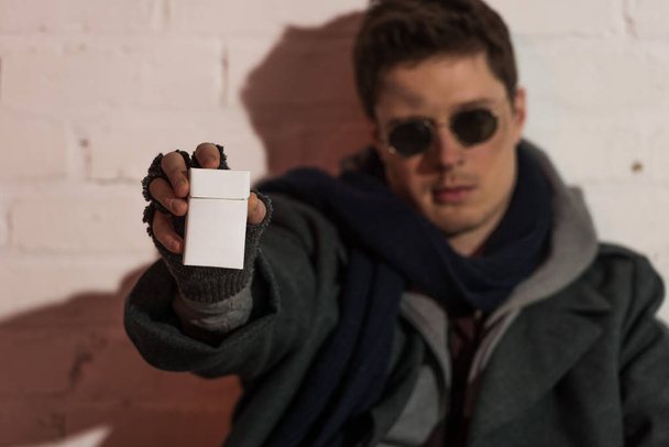 Selektivní fokus bezdomovec v rukavice bez prstů a tmavé brýle v natažené ruce držel krabičky cigaret - Fotografie, Obrázek