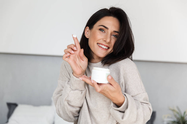 Image of smiling woman 30s holding jar with face cream in modern bright room - Fotó, kép
