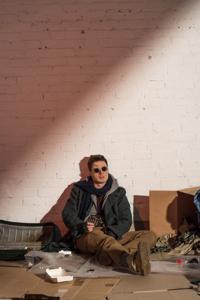 hajléktalan ember ül a fehér téglafal szemét körül - Fotó, kép