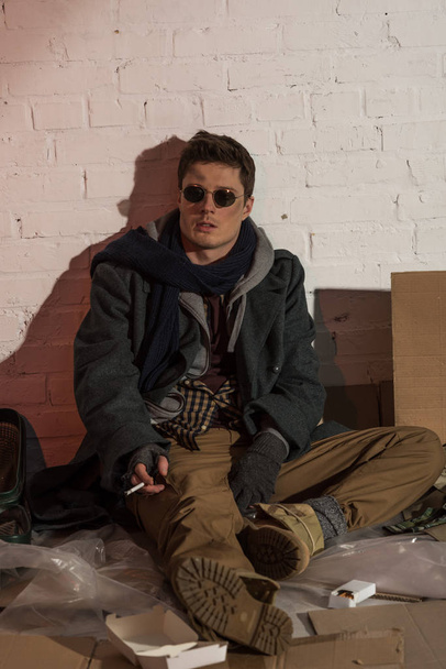 homeless man smoking while sitting on rubbish dumb by brick wall - Foto, Bild