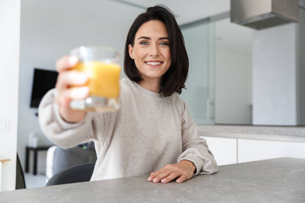 Image of beautiful woman 30s drinking orange juice while resting in bright modern room - Photo, Image