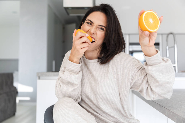 Imagen de mujer atractiva de 30 años sosteniendo dos piezas de naranja mientras está sentada en la sala de estar
 - Foto, imagen