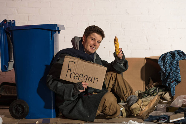 Lächelnder Obdachloser mit Maiskolben und Pappkarte mit "freegan" handgeschriebenem Schriftzug - Foto, Bild