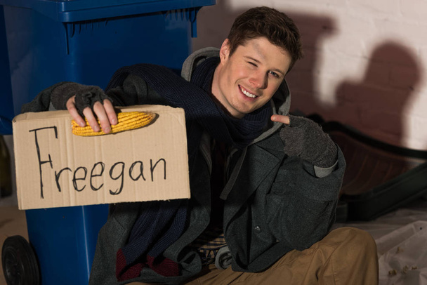 smiling homeless man holding corn cob and holding cardboard card with "freegan" inscription - 写真・画像