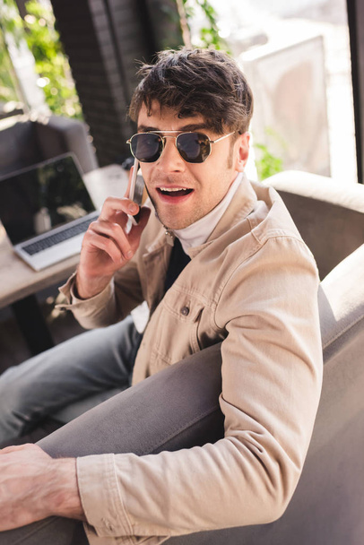 selective focus of man in sunglasses talking on smartphone near laptop in cafe - Photo, Image