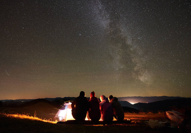 vue arrière d'un groupe d'amis randonneurs assis ensemble autour d'un feu de joie à côté d'une tente touristique lumineuse
 - Photo, image