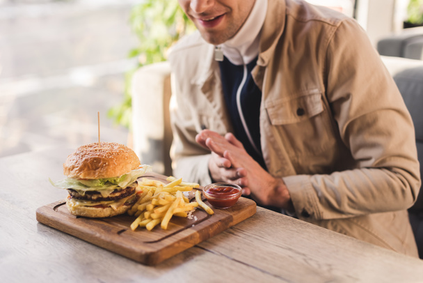 abgeschnittene Ansicht eines fröhlichen Mannes mit Blick auf leckere Burger und Pommes auf Schneidebrett im Café - Foto, Bild