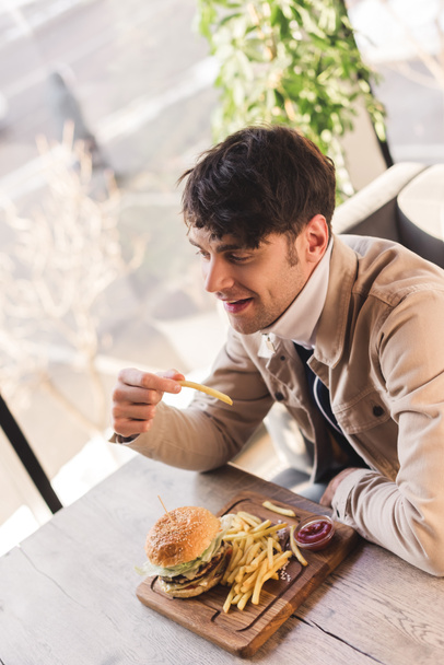 cheerful man holding french fry near tasty burger on cutting board in cafe - Fotó, kép