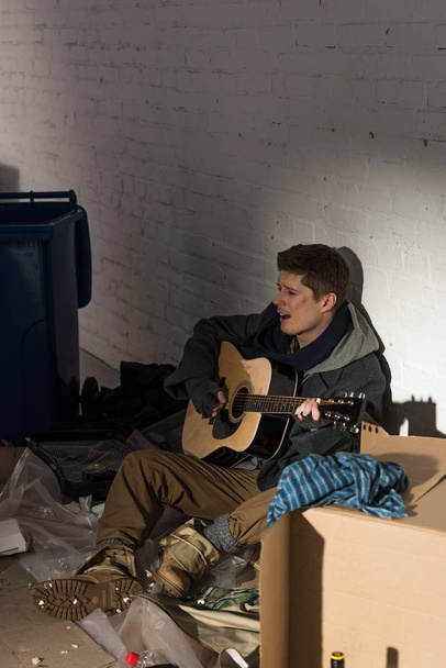 Obdachloser sitzt auf Pappe umgeben von Müll und spielt Gitarre - Foto, Bild