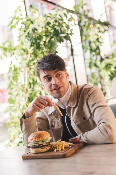 handsome young man holding french fry near tasty burger on cutting board in cafe - Photo, Image