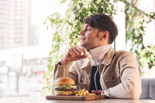cheerful young man holding french fry near tasty burger on cutting board in cafe - Фото, изображение