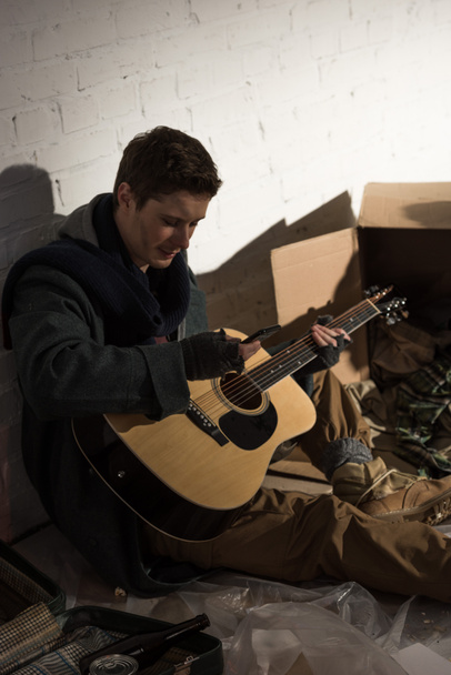 Бездомный держит гитару и использует смартфон, сидя на помойке
 - Фото, изображение