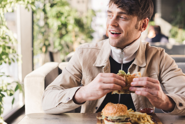 selective focus of happy young man holding tasty burger near french fries on cutting board in cafe - 写真・画像