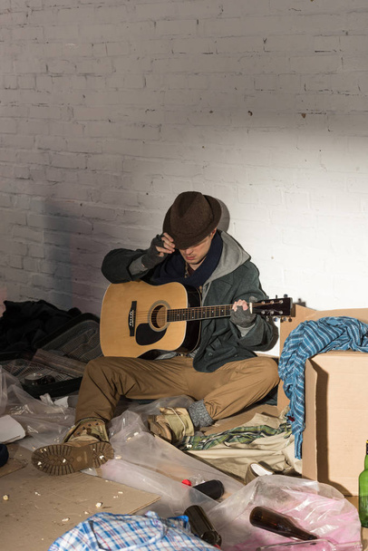 homeless man in hat and fingerless gloves holding guitar while sitting surrounded by rubbish - Foto, Bild