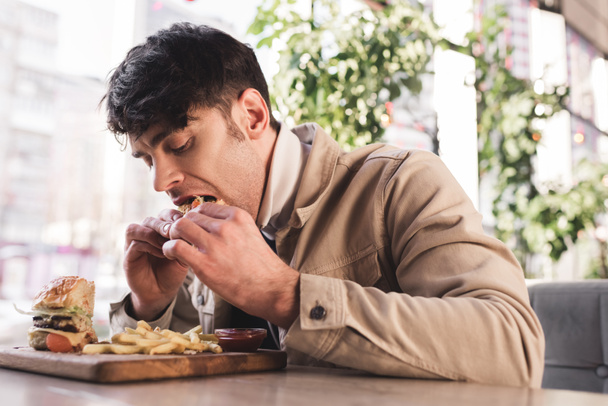 Junger Mann isst leckeren Hamburger in der Nähe von Pommes auf Schneidebrett in Café - Foto, Bild