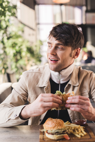 selective focus of smiling young man holding tasty hamburger near french fries on cutting board in cafe - Фото, зображення