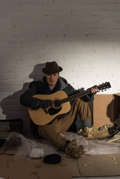 ホームレスの黒い服の男し、帽子はゴミ捨て場の上に座って、ギターを弾く - 写真・画像