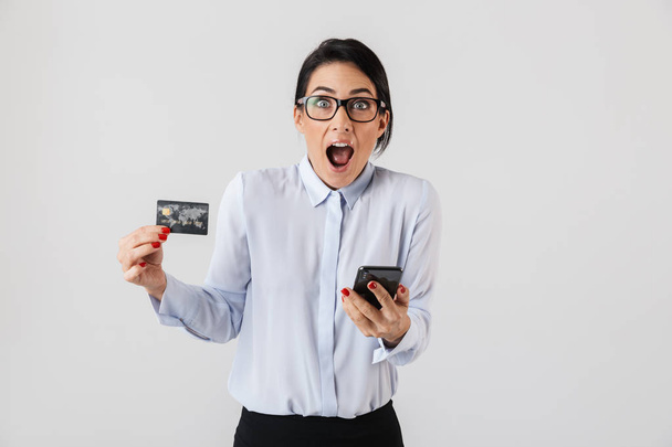 Image of european office woman wearing eyeglasses holding mobile phone and credit card isolated over white background - Photo, image