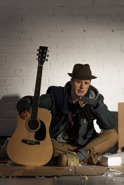 dakloze man gitaar zittend op karton omringd door vuil te houden - Foto, afbeelding