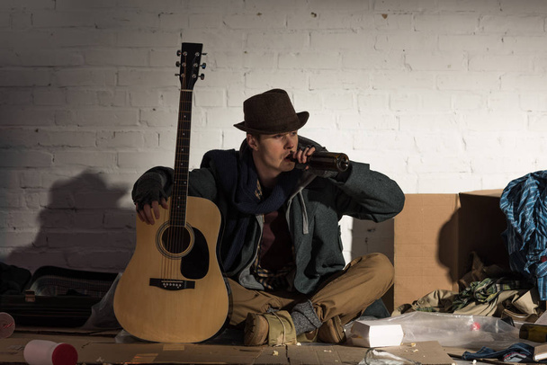 Obdachloser hält Gitarre und trinkt aus Alkoholflasche, während er auf Müllhalde sitzt - Foto, Bild