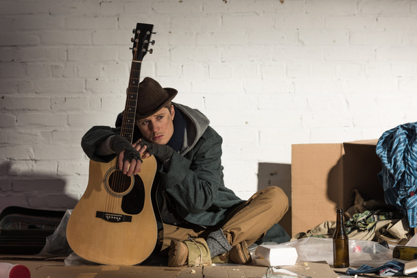 通りのゴミ捨て場に座ってギターに傾いているホームレスの男性を落ち込んでください。 - 写真・画像