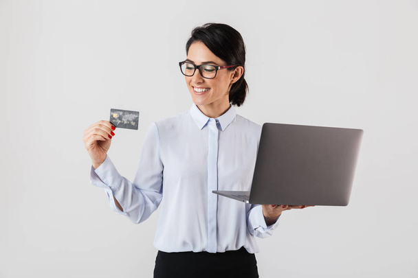 Portrait of adorable businesswoman wearing eyeglasses holding silver laptop and credit card in the office isolated over white background - Foto, Bild
