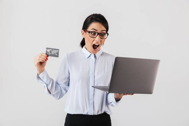 Portrait of european businesswoman wearing eyeglasses holding silver laptop and credit card in the office isolated over white background - 写真・画像