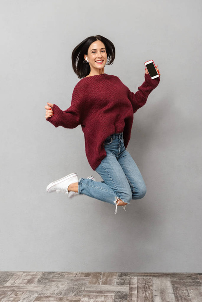 Full length portrait of excited woman 30s using earbud jumping and holding smartphone isolated over gray background - Foto, Imagem