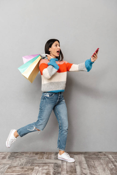 Portrait of emotional woman 30s running with colorful paper shopping bags and cell phone in hands isolated over gray background - Foto, Imagem