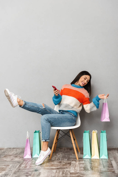 Image de femme brune 30s assis dans une chaise avec des sacs à provisions colorés et en utilisant un téléphone mobile isolé sur fond gris
 - Photo, image