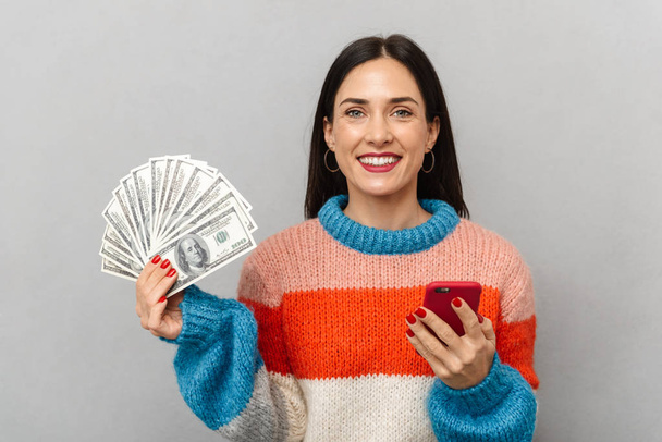 Фото чарівної жінки 30 років тримає купу грошей і смартфон ізольовано на сірому фоні
 - Фото, зображення
