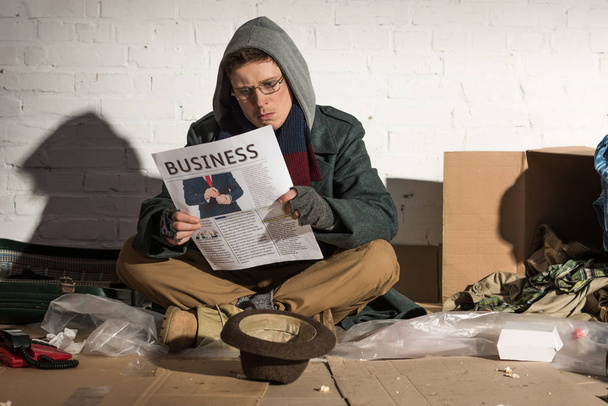 homeless beggar reading business newspaper while sitting by brick wall - Photo, Image