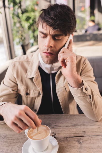 серйозний чоловік тримає ложку в чашці з капучино і розмовляє на смартфоні в кафе
 - Фото, зображення