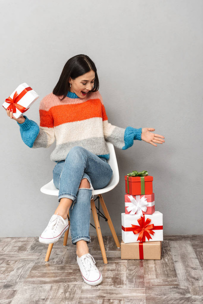 Photo of stylish woman 30s sitting in chair with present boxes isolated over gray background - Foto, Imagen