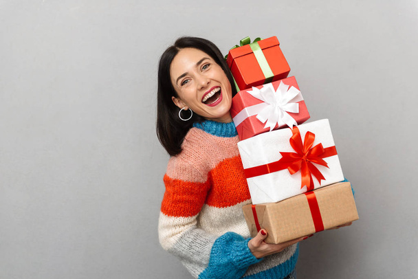 Foto de mujer positiva 30s sosteniendo racimo de cajas de regalo mientras está de pie aislado sobre fondo gris
 - Foto, imagen