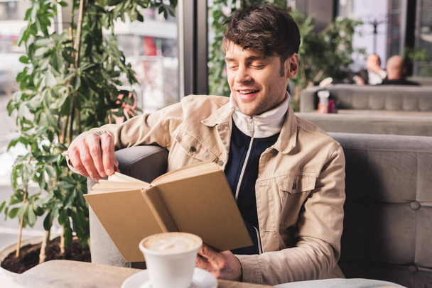 glücklicher Mann hält Buch neben Tasse mit Cappuccino im Café - Foto, Bild