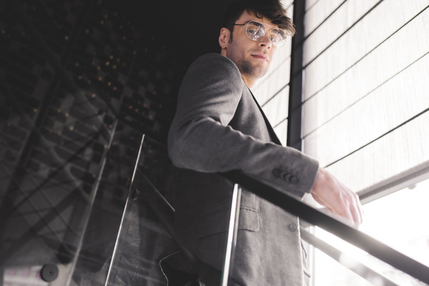 young stylish man in glasses standing on stairs and looking at camera - 写真・画像