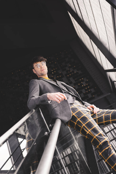 Stilvoller junger Mann mit Brille posiert auf der Treppe und blickt in die Kamera - Foto, Bild