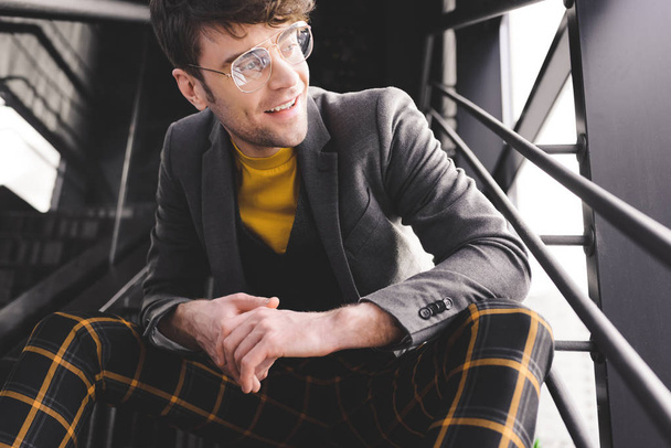 smiling stylish man in glasses sitting on stairs near windows - Фото, изображение
