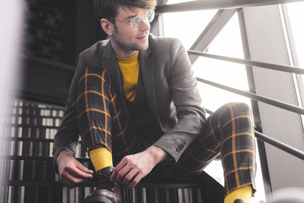 happy stylish man in glasses sitting on stairs and touching shoelaces while looking windows - Foto, afbeelding