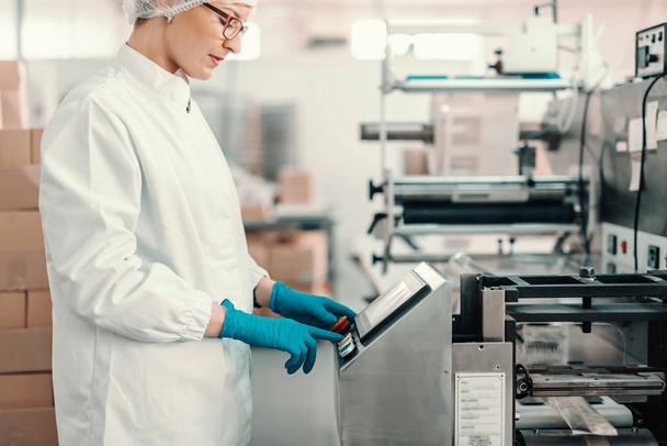 Joven empleada en uniforme estéril y guantes de goma azul encendiendo la máquina de embalaje mientras está de pie en la fábrica de alimentos
. - Foto, imagen