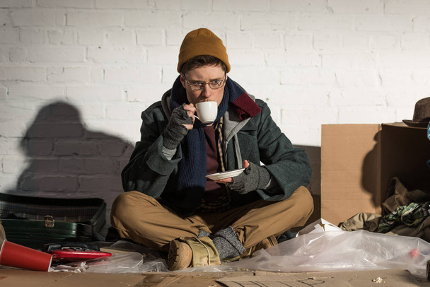 bezdomny człowiek picie kawy siedząc przez biały mur na wysypisku śmieci - Zdjęcie, obraz